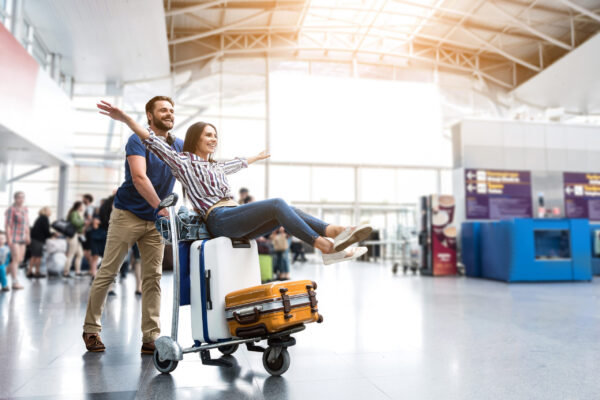 I believe I can fly. Cheerful woman is sitting on baggage and man pulling cart forward. They looking ahead with joyous smile. Copy space on right side